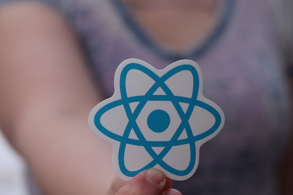 A person holds a small sticker with a blue atomic model symbol in front of their chest, which features a central circle surrounded by interconnected elliptical orbits. The background shows a blurred v