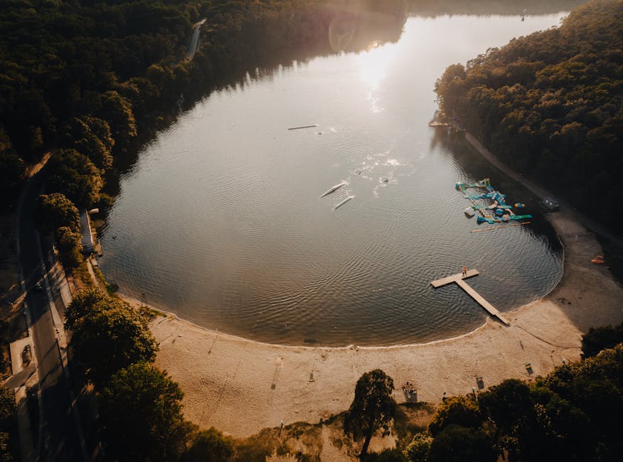 An aerial view of a large body of water surrounded by dense green trees and vegetation under a partly cloudy sky with the sun low on the horizon. Nearby, a small pier or boathouse extends into the wat